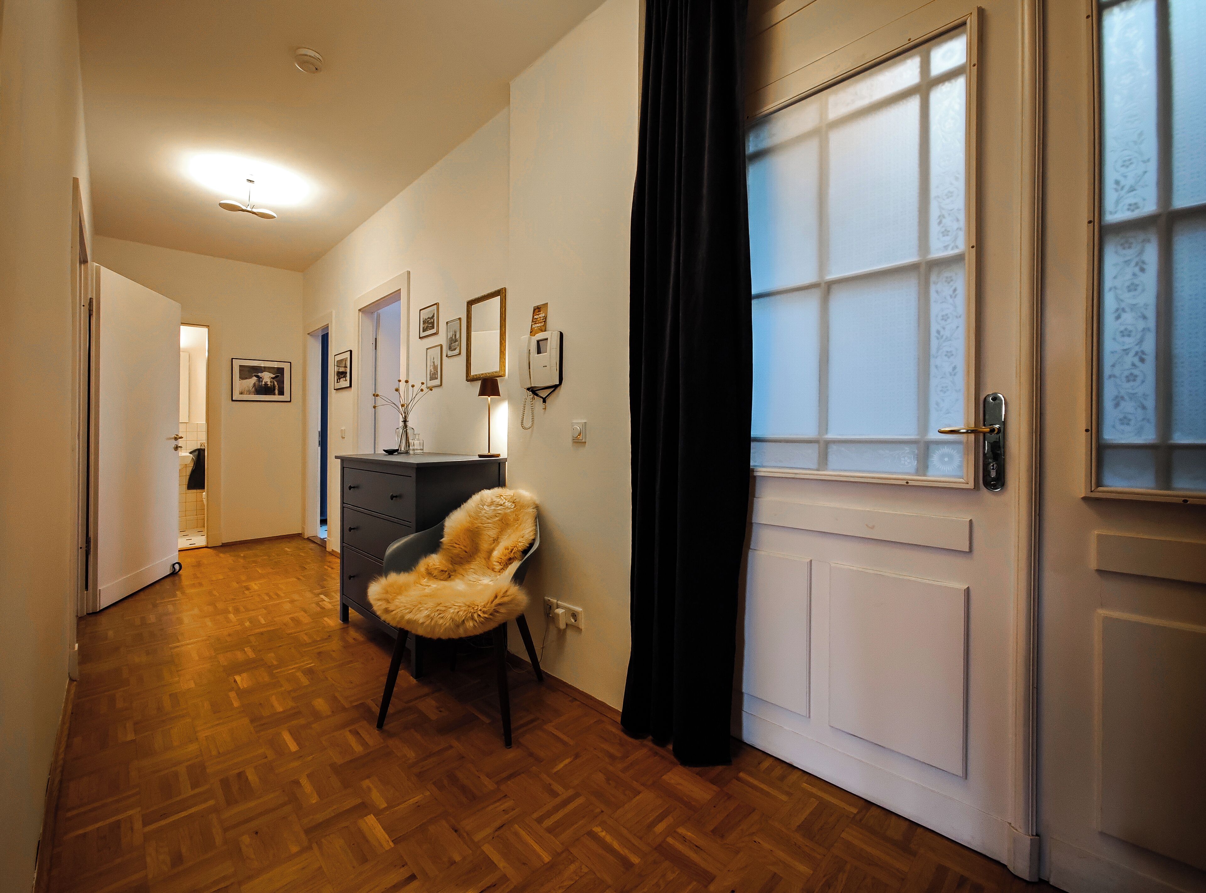 Un couloir doté d’un plancher en bois, d’une chaise, d’une commode et d’une porte avec des panneaux en verre dépoli.