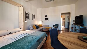 A modern, well-lit room with a bed, sofa, and dining table. There are framed pictures on the walls and a television on a stand.