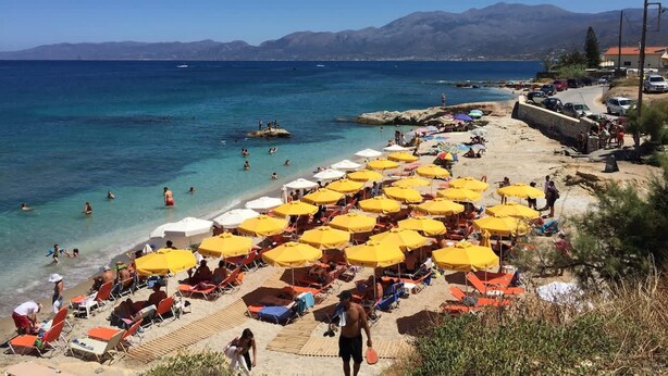 Een strand met tal van gele parasols en rode ligbedden, een heldere blauwe zee en bergen op de achtergrond.