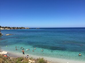Una platja amb aigua turquesa cristal·lina, gent banyant-se i prenent el sol, i roques al fons.