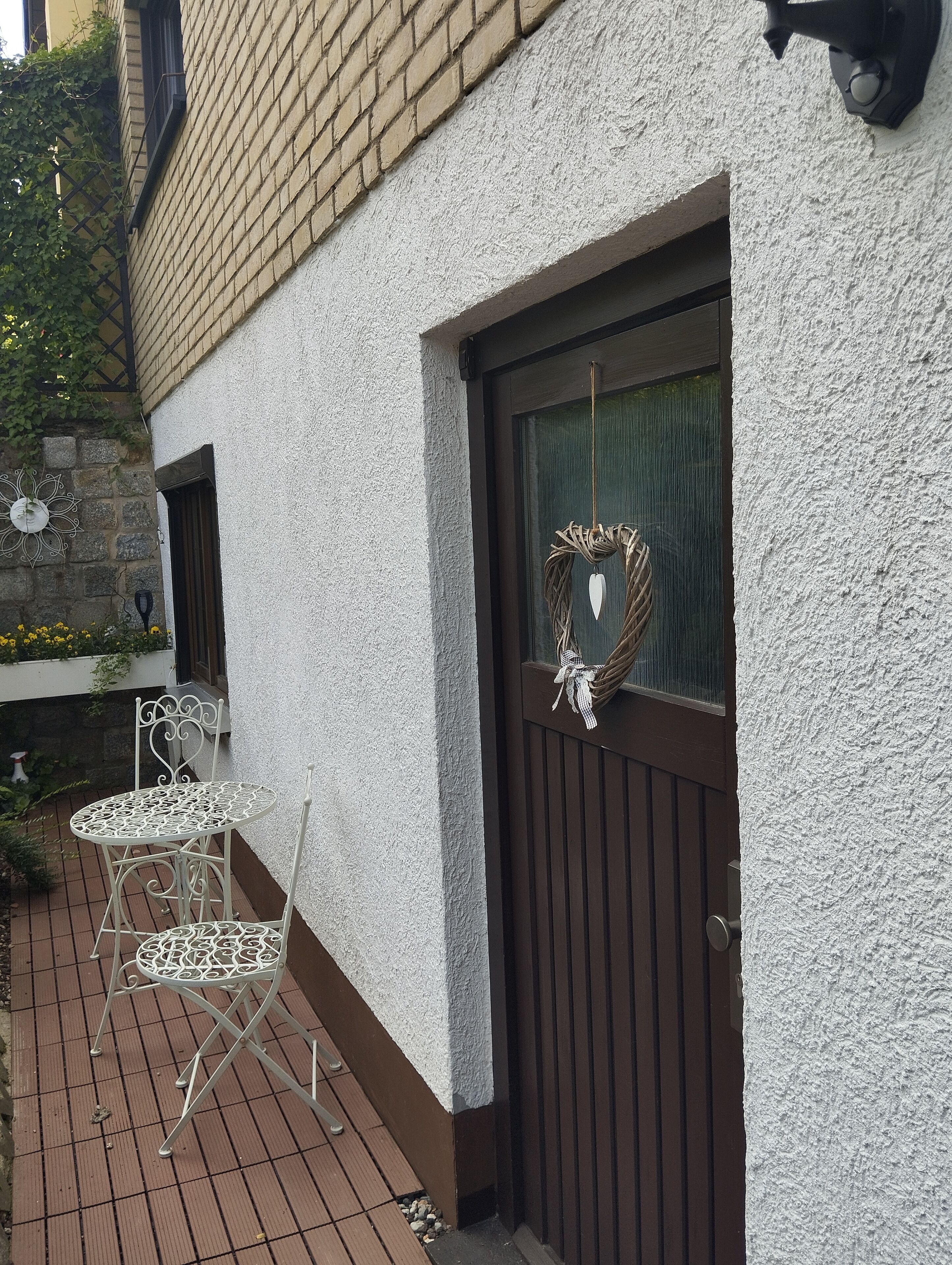 Un mur extérieur blanc avec un revêtement texturé, une porte marron ornée d’une guirlande décorative, ainsi qu’une petite table et une chaise en métal blanc, le tout installé sur une terrasse en briques.