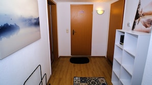 A hallway with a wooden door, a white wall with a painting, a black mat, and a white shelf with a picture.