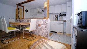 A compact kitchen with white cabinets, a microwave, and a coffee maker. There is a dining area with a table and chairs, and a wicker chair.