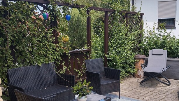 An outdoor patio with wicker furniture, a small table with a plant, and a pergola.