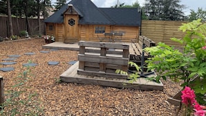 Una cabaña de madera con techo inclinado, una terraza de madera y un jardín con piedras y plantas.