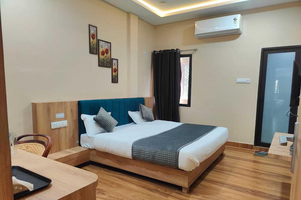 A hotel room with a bed, wooden floor, a desk, and an air conditioning unit.