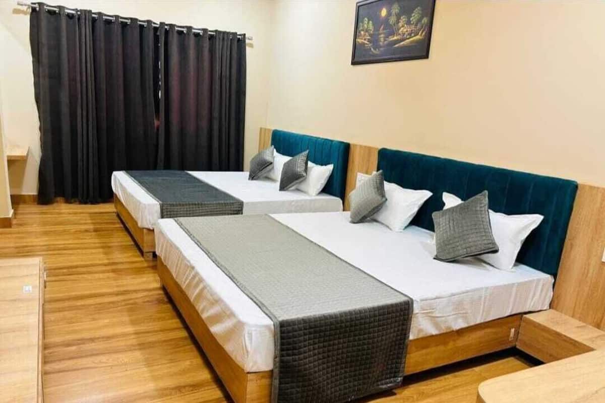 A room with two beds, each with a blue headboard and white bedding. There is a wooden floor and a black curtain on the window.