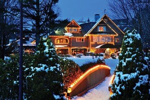 A large, multi-story wooden house with a snow-covered driveway and a lit bridge.