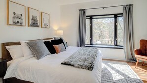 Une chambre à coucher comprenant un lit, une table de chevet, une chaise, une fenêtre avec des rideaux et des œuvres d’art murales.