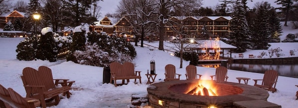 A snow-covered outdoor area with a fire pit and Adirondack chairs, a pond, and lodge buildings in the background. - Condo 3B/3BA + Amenity Pass w/ Hot Tub + Pool. (Blowing Rock)