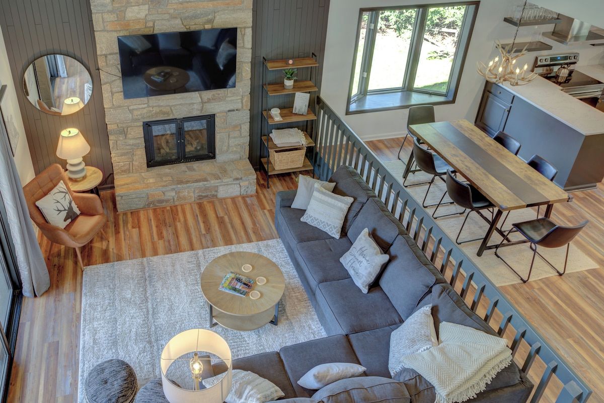 A modern living room with a stone fireplace, a television mounted above it, and a dining area with a wooden table and chairs.
