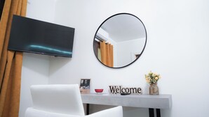 Ein modernes Hotelzimmer mit einem Wand-Fernseher, einem runden Spiegel, einem weißen Stuhl, einem kleinen Tisch mit einem 'Welcome'-Schild, einer roten Schale und einer Vase mit Blumen.