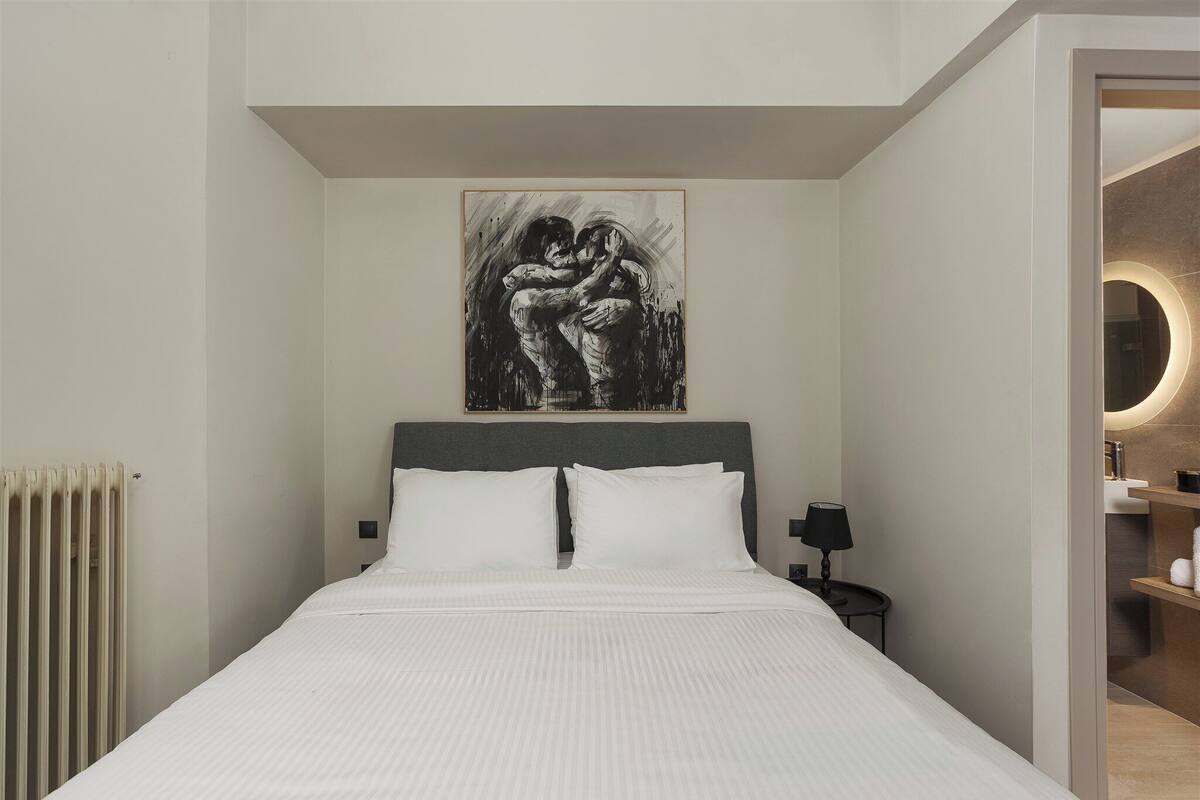 Ein Schlafzimmer mit einem großen Bett, einem Kopfteil, einem Nachttisch mit Lampe und einem an der Wand hängenden Kunstwerk.