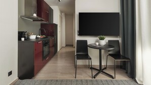 A modern kitchen with dark cabinetry, a built-in oven, and a stainless steel range hood. There is a dining area with a round table and two black chairs, and a large flat-screen TV mounted on the wall. - Anvy Stylish Suites Ioannina (Ioannina)