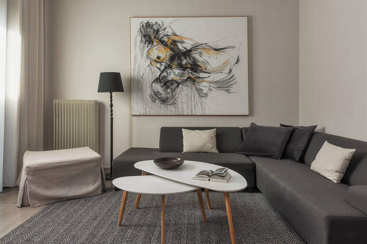 Ein modernes Wohnzimmer mit einem grauen Sofa, einem weißen Couchtisch und einer schwarzen Stehlampe. Ein großes abstraktes Gemälde hängt an der Wand, und ein Heizkörper ist zu sehen.