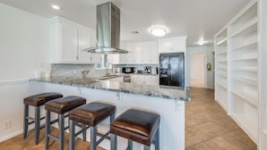 A modern kitchen with white cabinets, a central island with stools, stainless steel appliances, and a tiled floor.