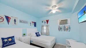 Ein Schlafzimmer mit zwei Betten, einem Deckenventilator und Wanddekorationen, darunter Flaggen und ein Schild mit der Aufschrift 'BEACH'.
