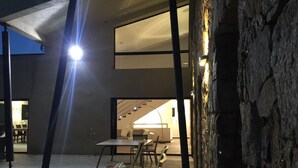 Ein Essbereich im Freien mit Tischen und Stühlen, einer Steinmauer und einer hellen Lichtquelle.