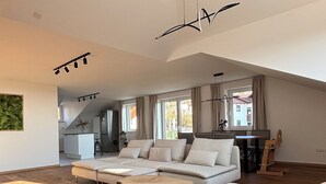 A spacious living area with a large sofa, a smaller sofa, and a wooden coffee table. There is a kitchen area in the background with a refrigerator and a dining table.