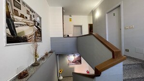 Un couloir étroit avec un escalier en bois, un tableau encadré au mur et une petite fenêtre au fond.