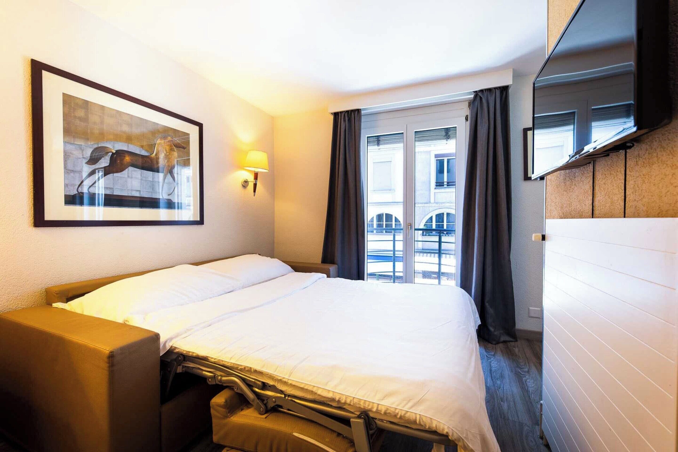 Ein Hotelzimmer mit Bett, Schreibtisch, Stuhl, einem gerahmten Bild an der Wand, einem Fenster mit Vorhängen und einem an der Wand befestigten Flachbildfernseher.