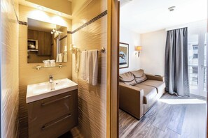 Un baño moderno con tocador de vidrio, un espejo grande y un armario de madera. Junto a él, un dormitorio con sofá y ventana amplia.