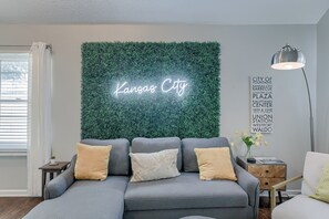 Una sala de estar moderna con un sofá gris, cojines decorativos, un letrero de neón que dice "Kansas City" y un cartel en la pared que enumera atracciones locales.