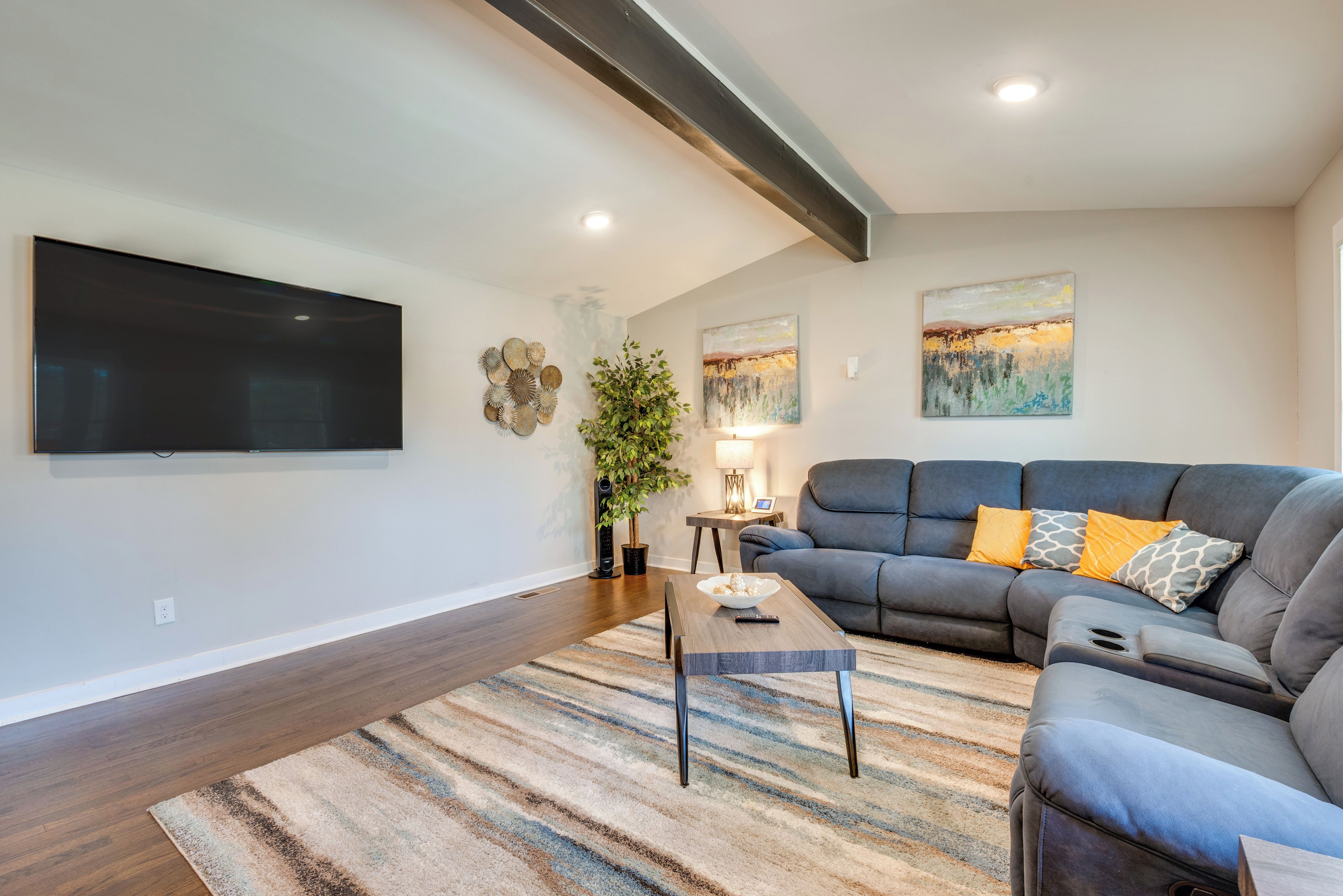 A modern living room with a large flat-screen TV, a grey sectional sofa with yellow and grey cushions, a wooden coffee table, and a striped rug.