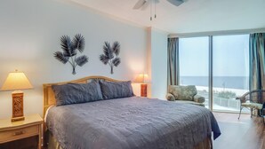 Un dormitorio con una cama grande, dos lámparas de pared, un ventilador de techo y vistas al mar a través de amplios ventanales.