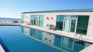 Una piscina con vistas despejadas al océano, un edificio con amplios ventanales y algunas tumbonas.