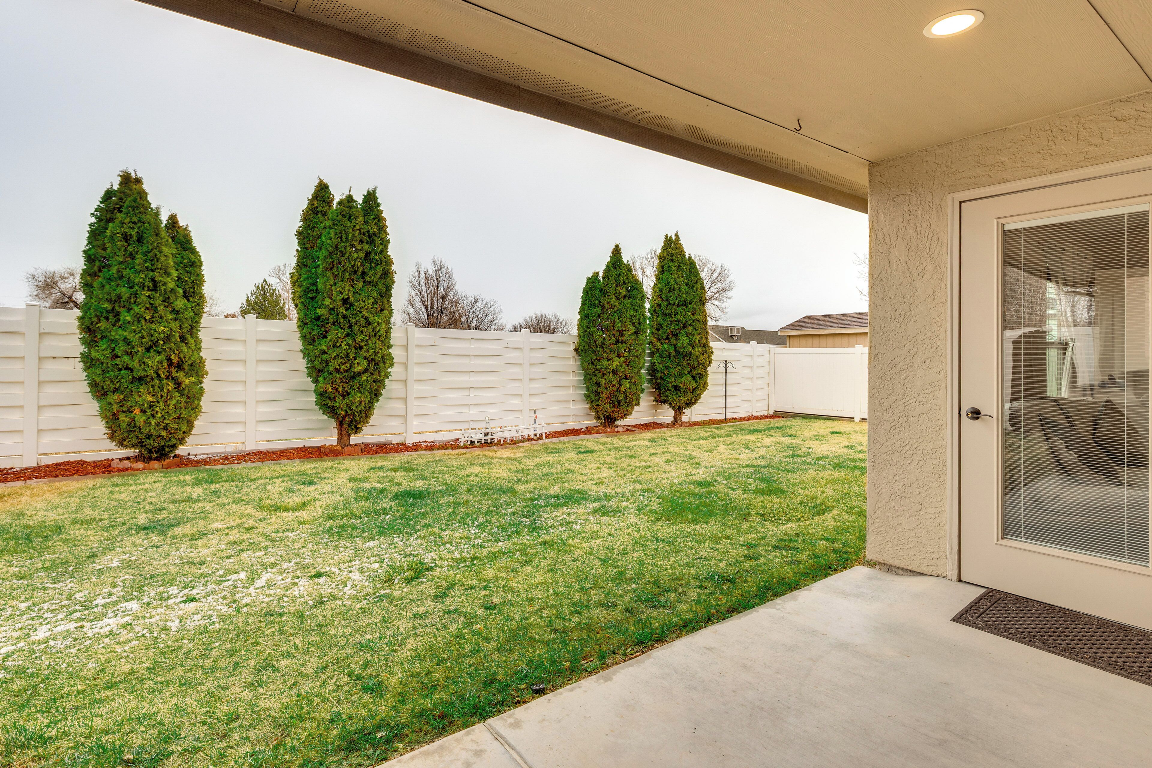 A covered patio area with a concrete floor, white fence, and three evergreen trees.