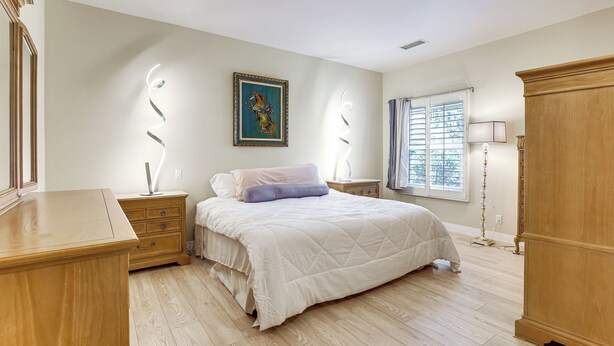 Une chambre avec un grand lit, une commode en bois, des tables de chevet, une petite table de nuit, une fenêtre avec des stores et un tableau accroché au mur.