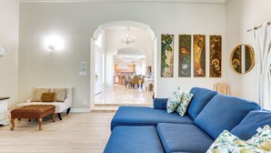 Un salon avec un canapé bleu, un pouf marron et un divan blanc. Les murs sont ornés de panneaux décoratifs et un miroir rond y est accroché.