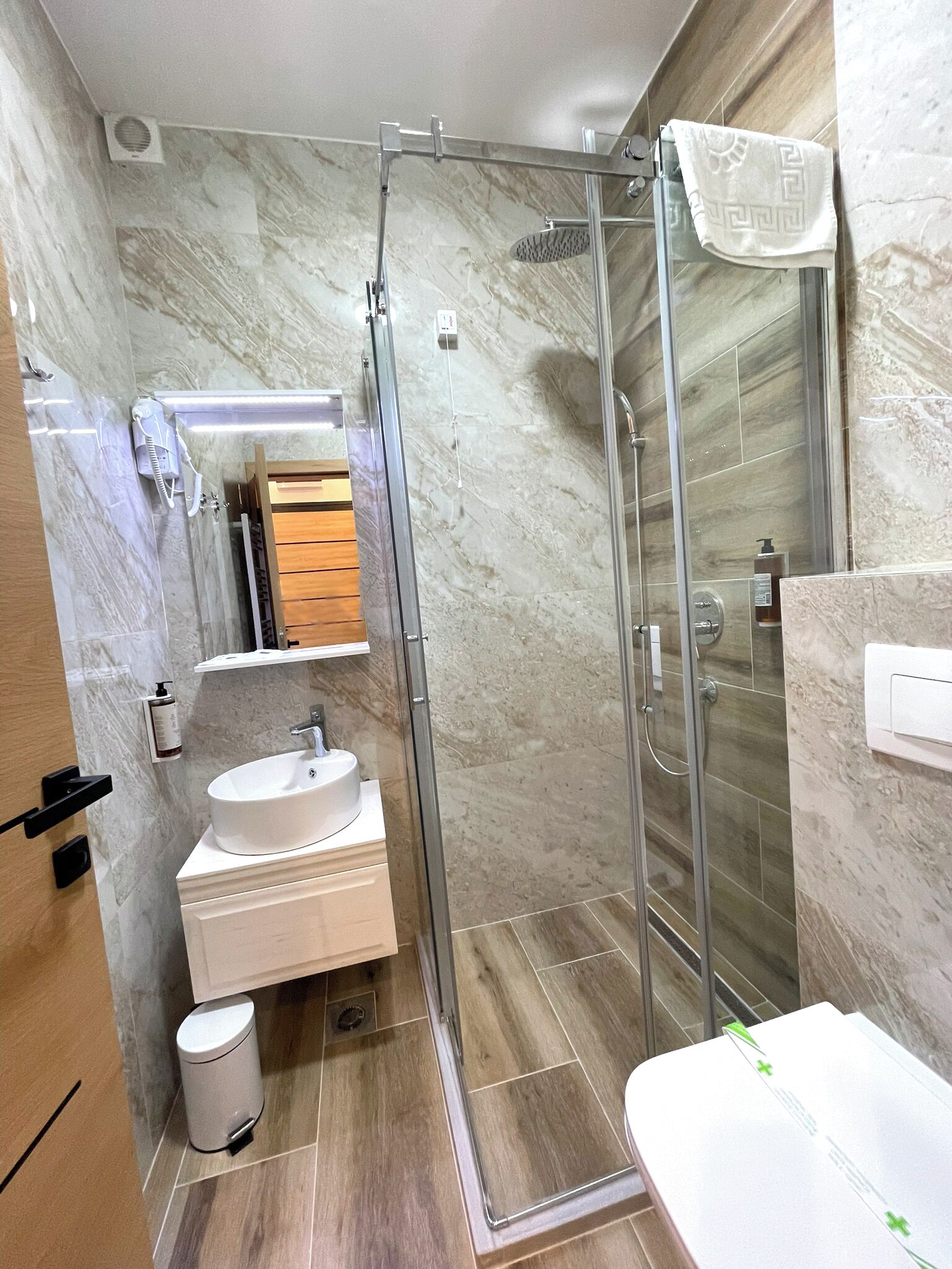 Een moderne badkamer met een douchecabine van glas, een wit wastafeltje en een houten opbergmeubel.