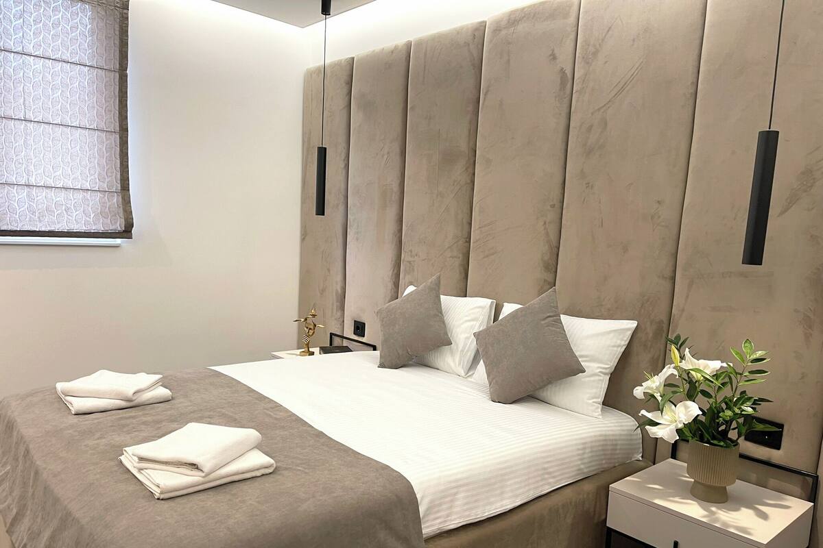 Ein modernes Schlafzimmer mit einem großen Bett, einem Nachttisch mit Blumenvase und einem Fenster mit Jalousien.