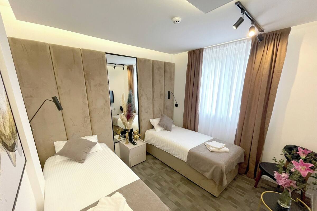 Ein Hotelzimmer mit zwei Betten, einem Spiegel, einem Nachttisch, einem kleinen Tisch mit Blumenvase und einem Fenster mit Vorhängen.