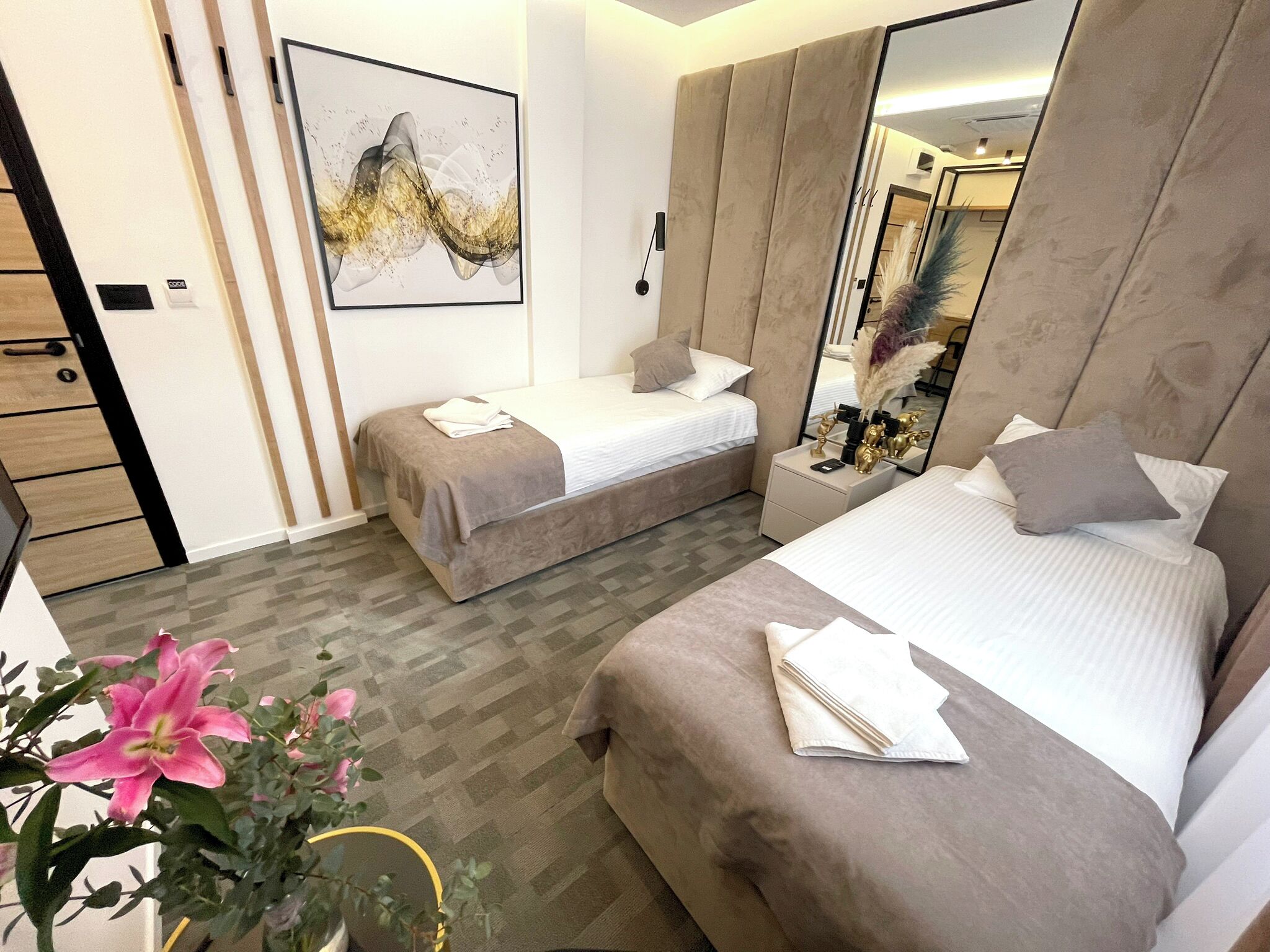 A modern hotel room with two beds, a large mirror, a decorative wall piece, and a potted plant.