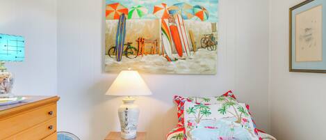 A bedroom with a beach-themed bedspread, a painting of beach umbrellas and surfboards, a bedside lamp, and a wooden dresser.