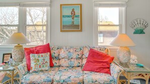 Une pièce avec un canapé à motifs floraux, deux lampes, un tableau encadré représentant une personne sur une plage et un éventail décoratif accroché au mur.