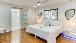 Ein Schlafzimmer mit einem Bett, einem Deckenventilator, einem Fenster mit Jalousien und einem an der Wand befestigten Spiegel.