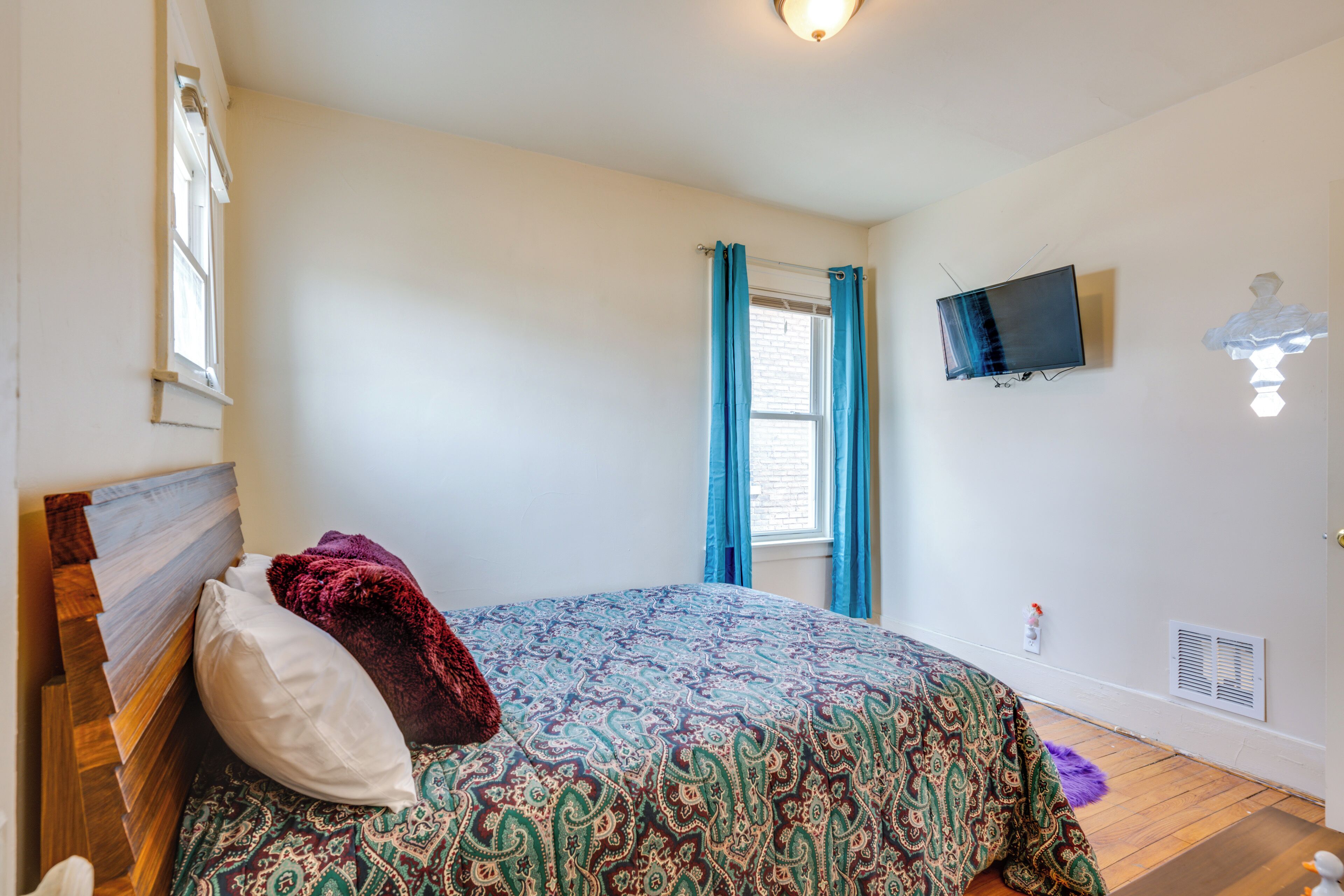 Un dormitorio con cama, ventana con cortinas, televisor montado y lámpara de pared.