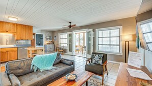 A spacious living area with a sofa, coffee table, and a dining table. There is a kitchen area with wooden cabinets and a refrigerator. The room has large windows with blinds and a ceiling fan. - Relax & Refresh: Waterfront Cottage in Panacea (Panacea)