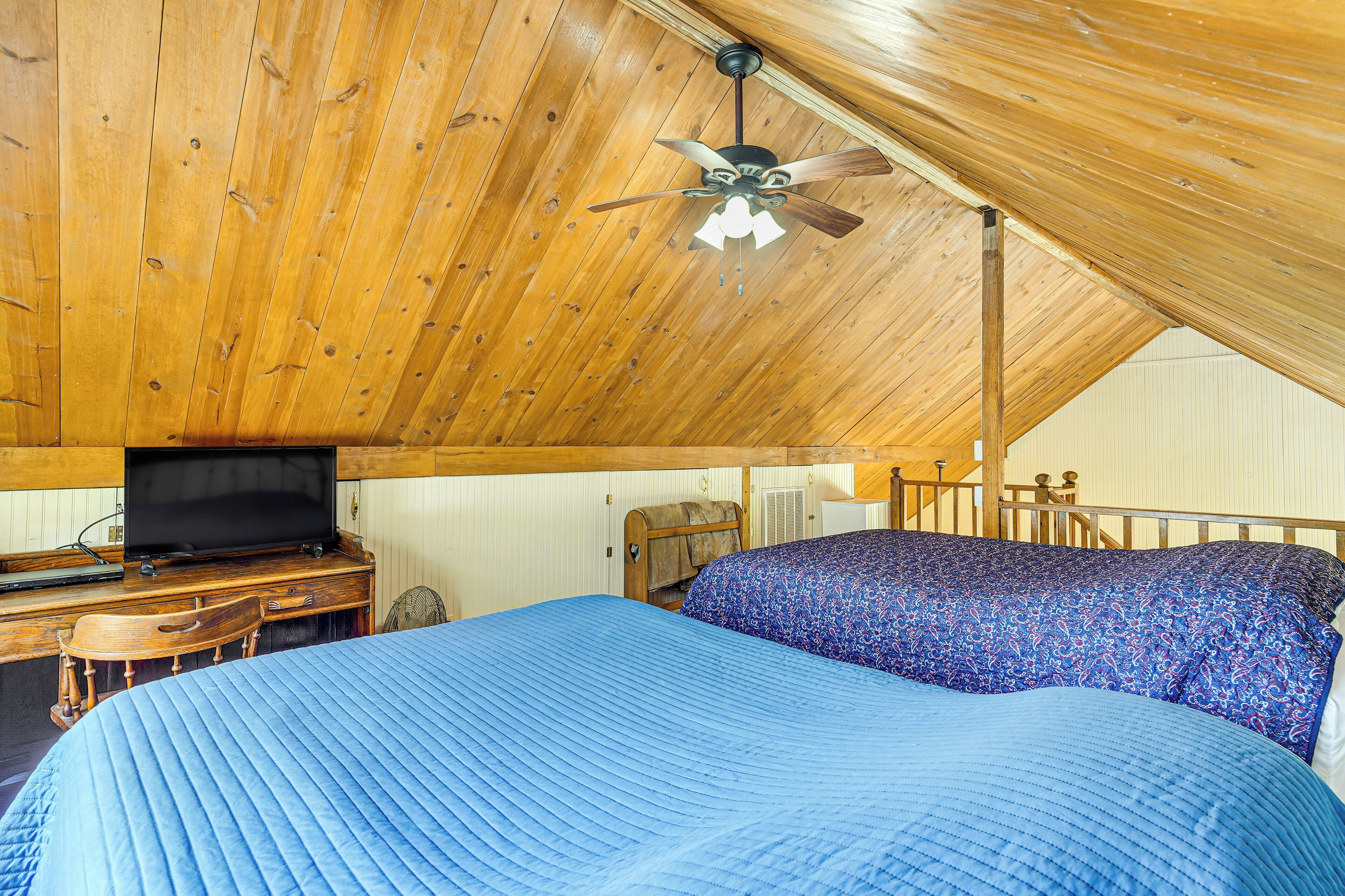 Quarto com teto de madeira, cama com roupa de cama azul, escrivaninha com televisão e uma cadeira.