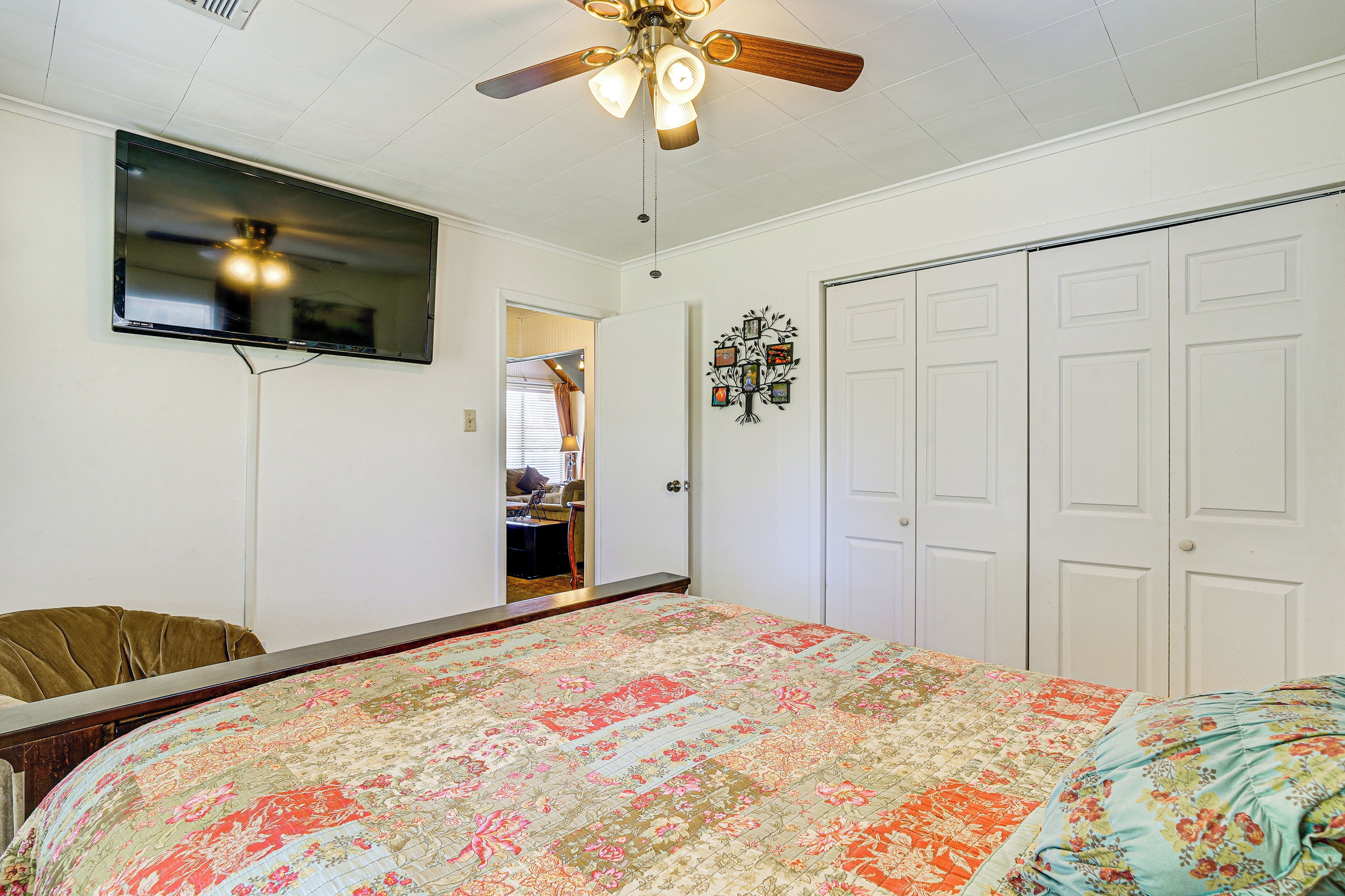A bedroom with a large bed, a ceiling fan, a TV mounted on the wall, and a door leading to another room.