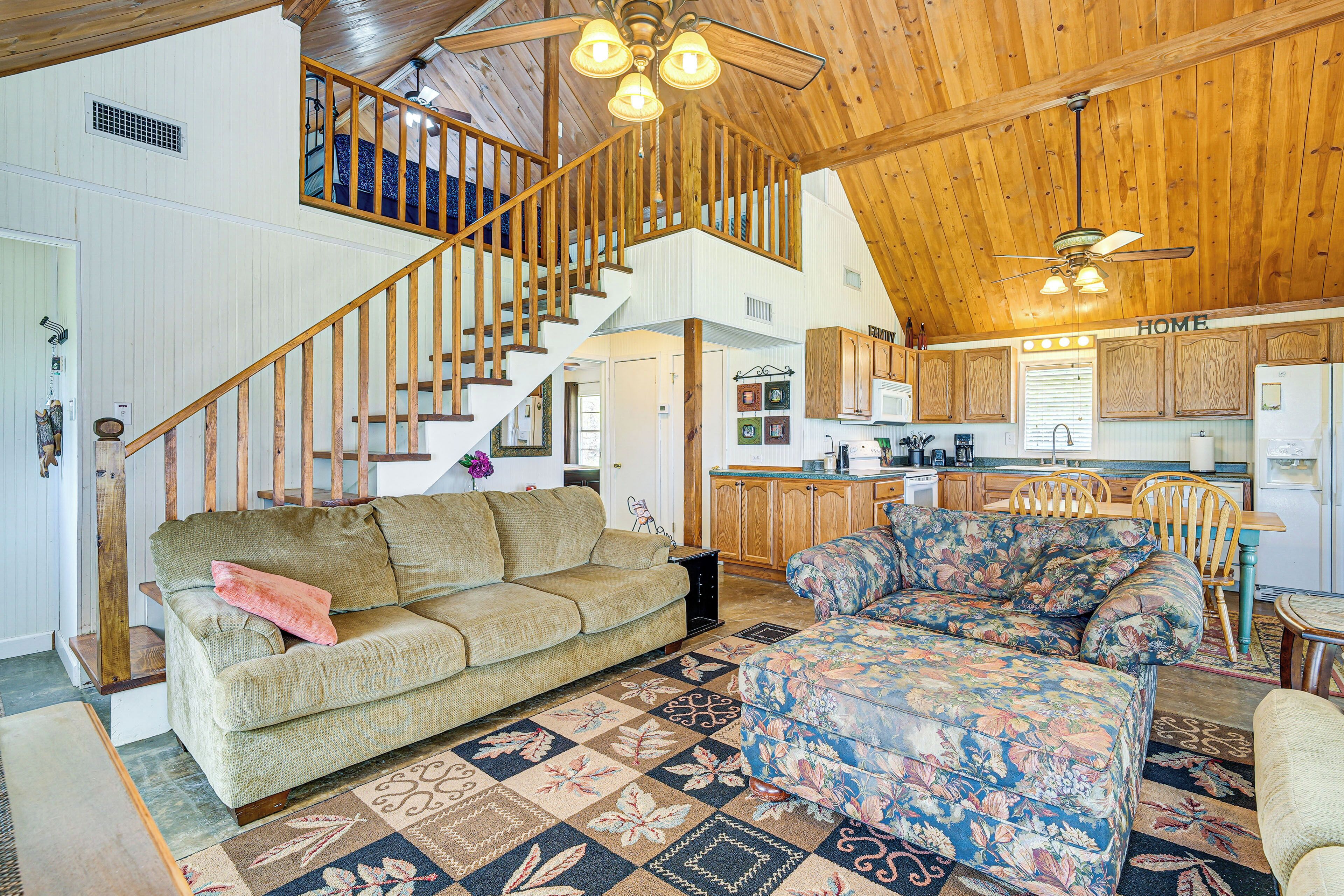 A spacious living area with a wooden staircase, a sofa, a floral ottoman, and a kitchen in the background.