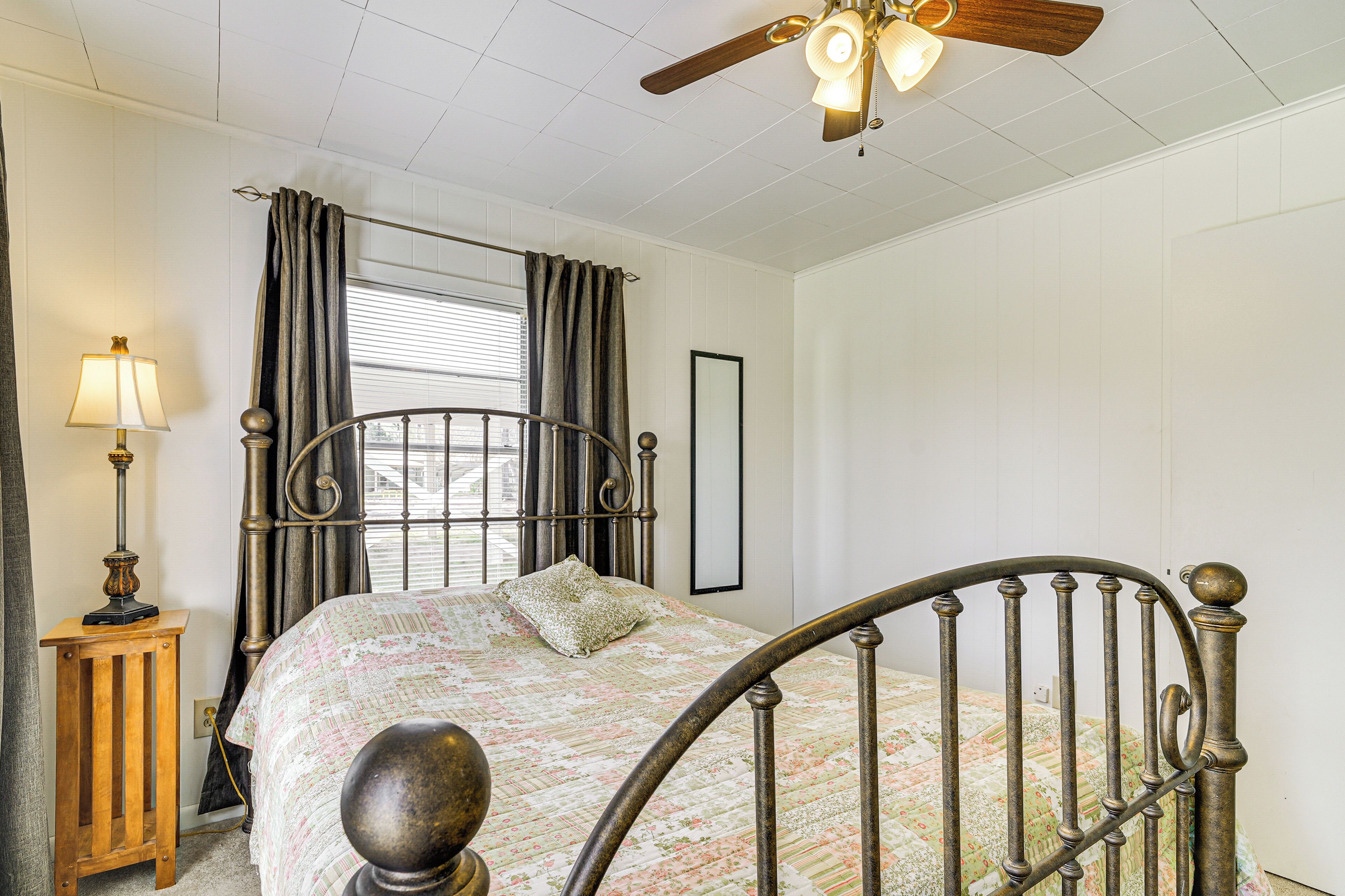 A bedroom with a bed, a nightstand, a lamp, a window with curtains, and a ceiling fan.