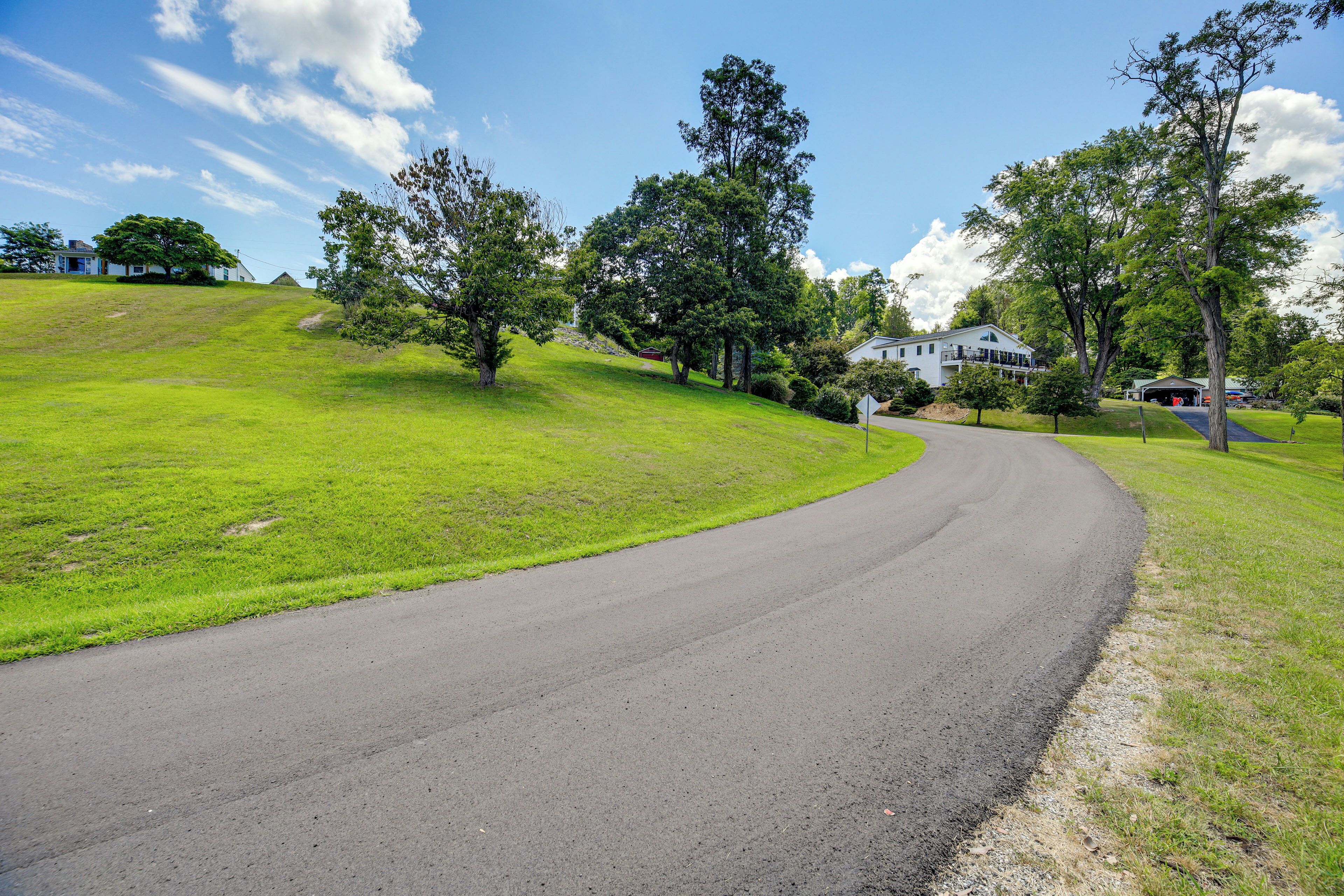 A curving road with a grassy hill, trees, and a white building with a garage in the background.