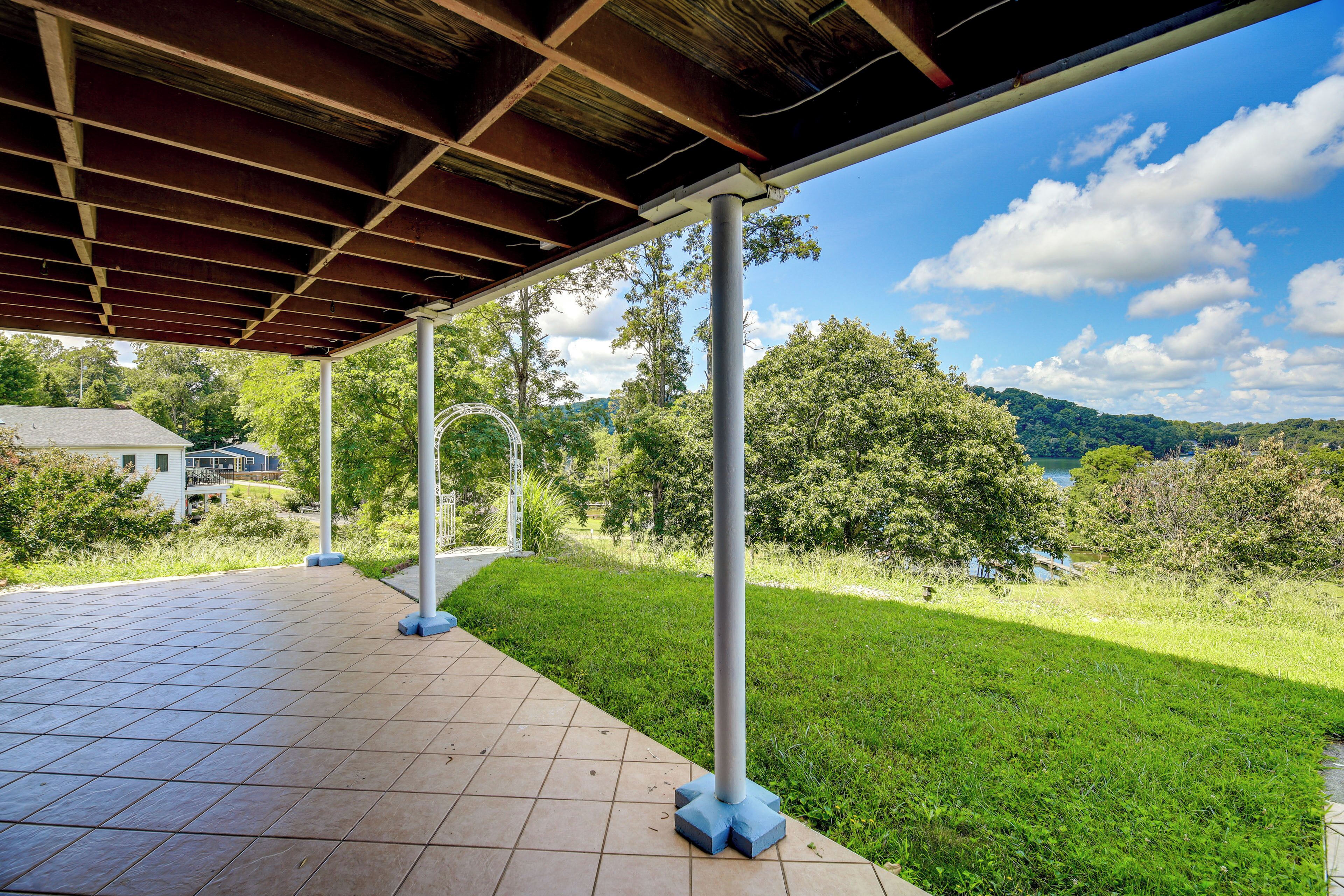 Un patio techado al aire libre con suelo de baldosas, columnas de metal y vistas a un lago y colinas.