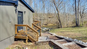 Un chalet rustique avec un escalier en bois menant à une terrasse, entouré d’un paysage naturel avec un ruisseau et des arbres.