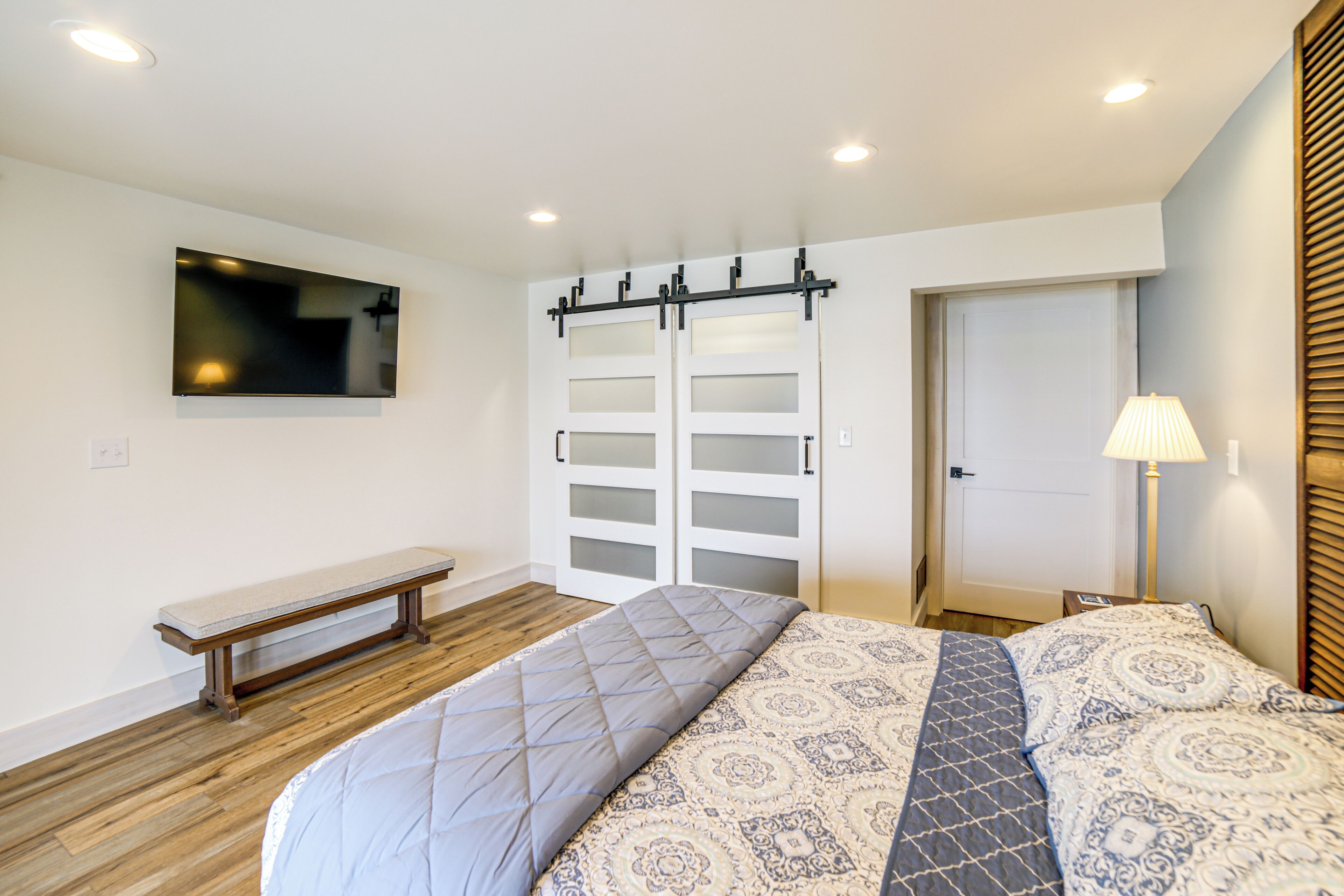A bedroom with a bed, a bench, a TV mounted on the wall, a door with frosted glass panels, and a lamp on a bedside table.